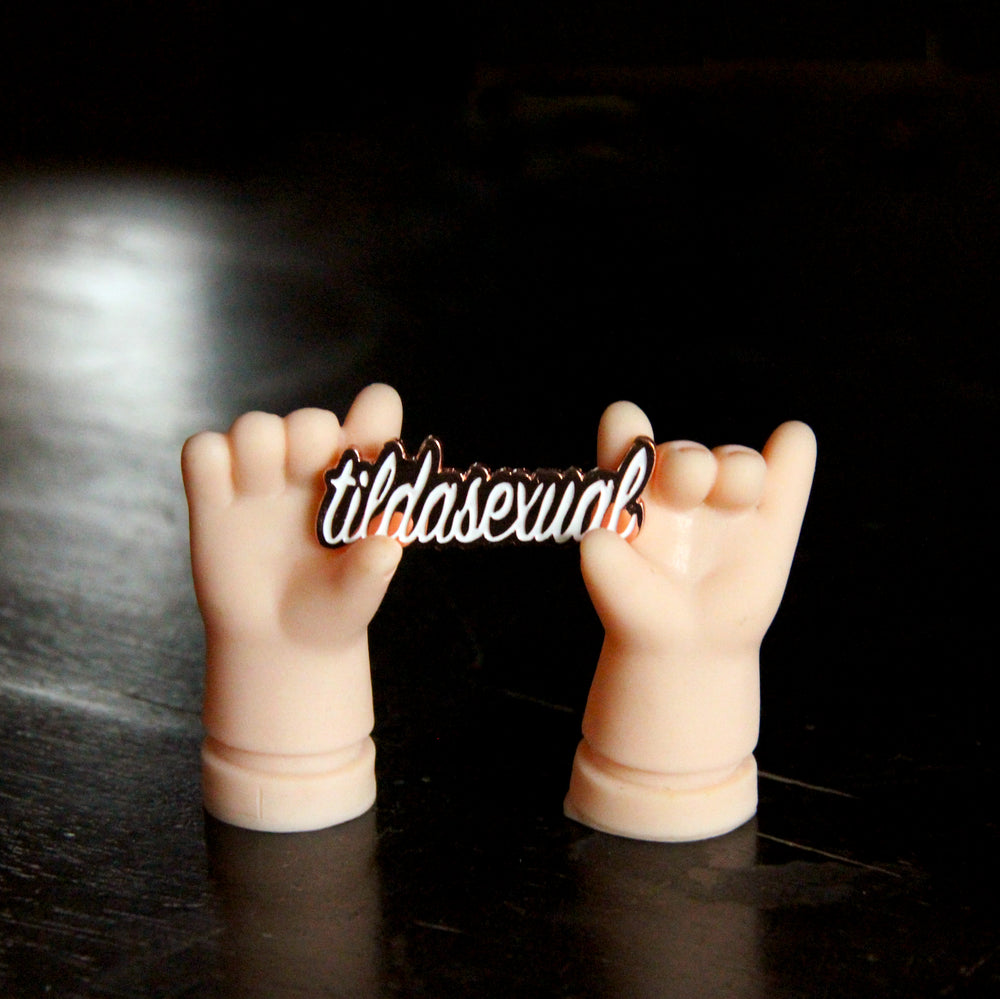 A pair of plastic doll hands set upon a black table hold up a tildasexual pin.
