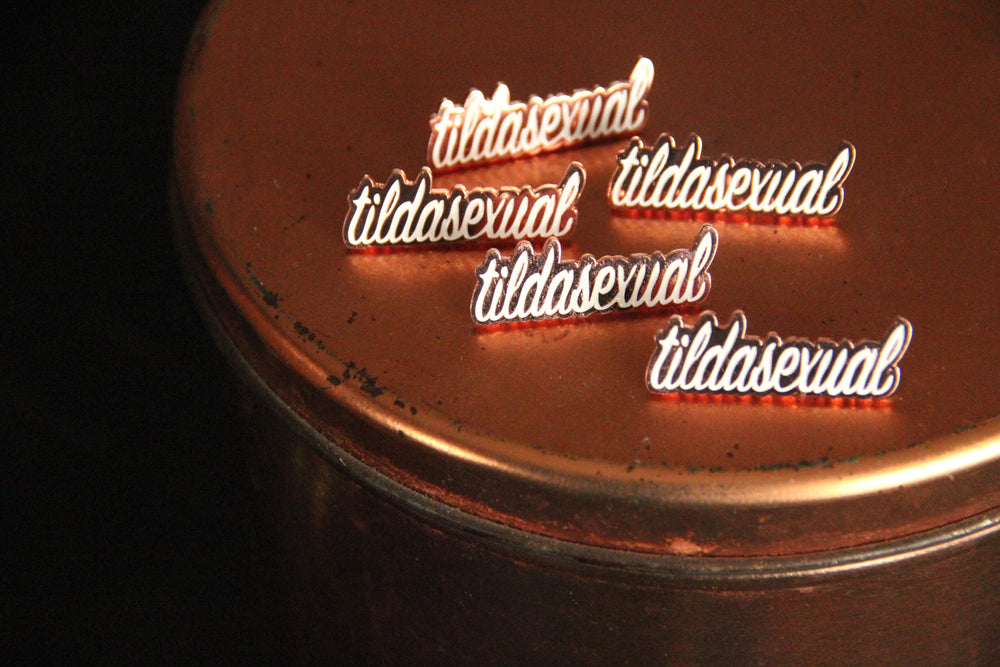 Four copper and white tildasexual pins are placed on the lid of a rustic copper tin.