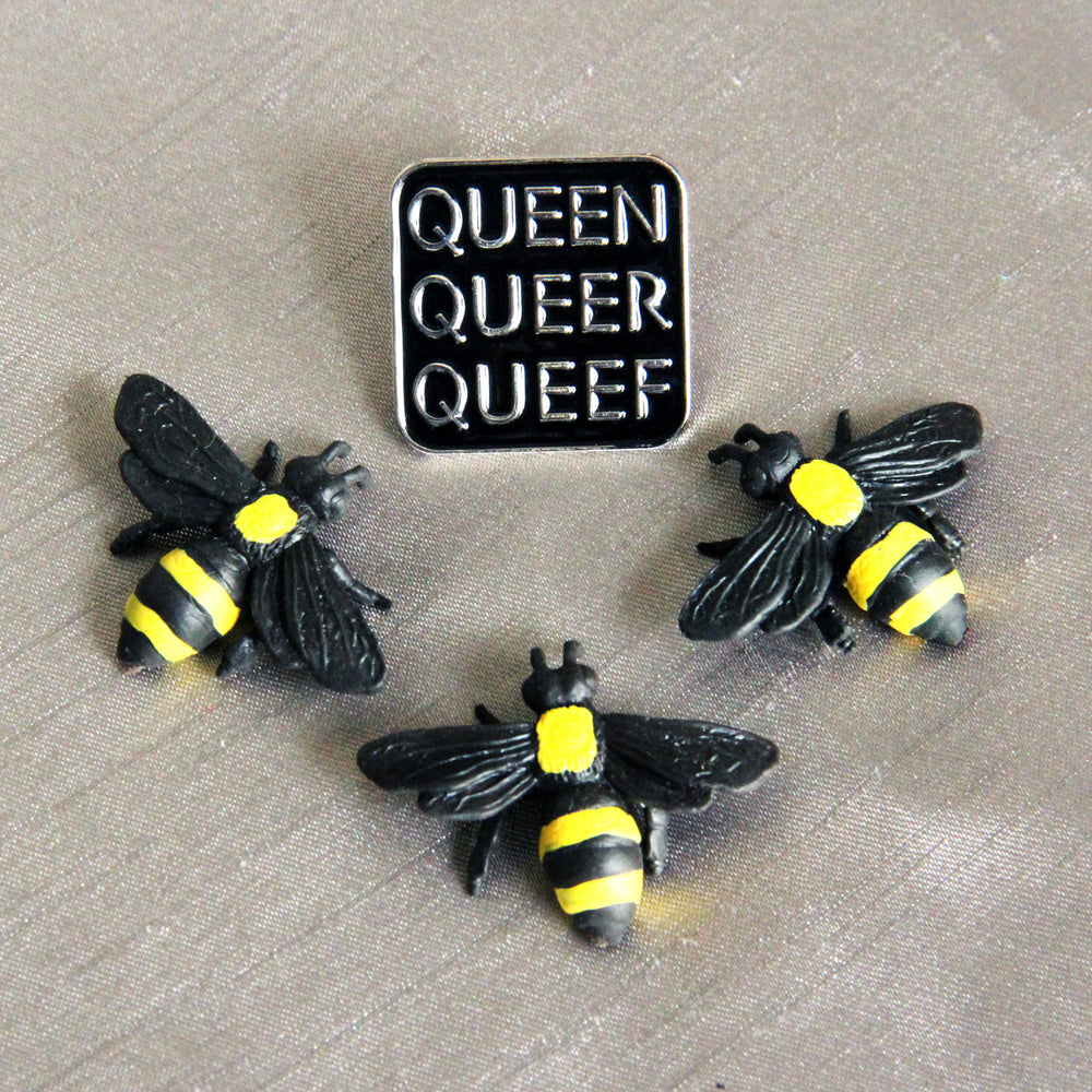 A trio of plastic toy bees stand before a Q-words pin on a silver-toned fabric background.