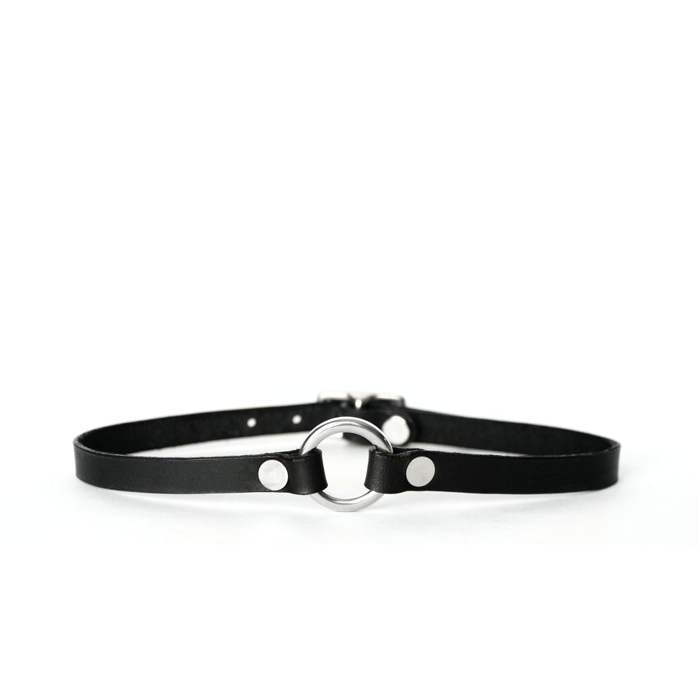 A thin black leather choker with a small silver o-ring at center front sits on a white surface with a white background.