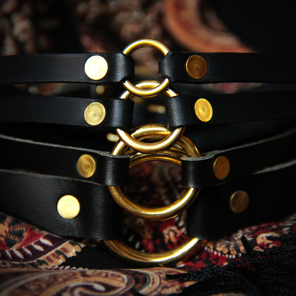 Four black chokers with brass hardware are tidily stacked from largest o-ring size to smallest on a paisley background.