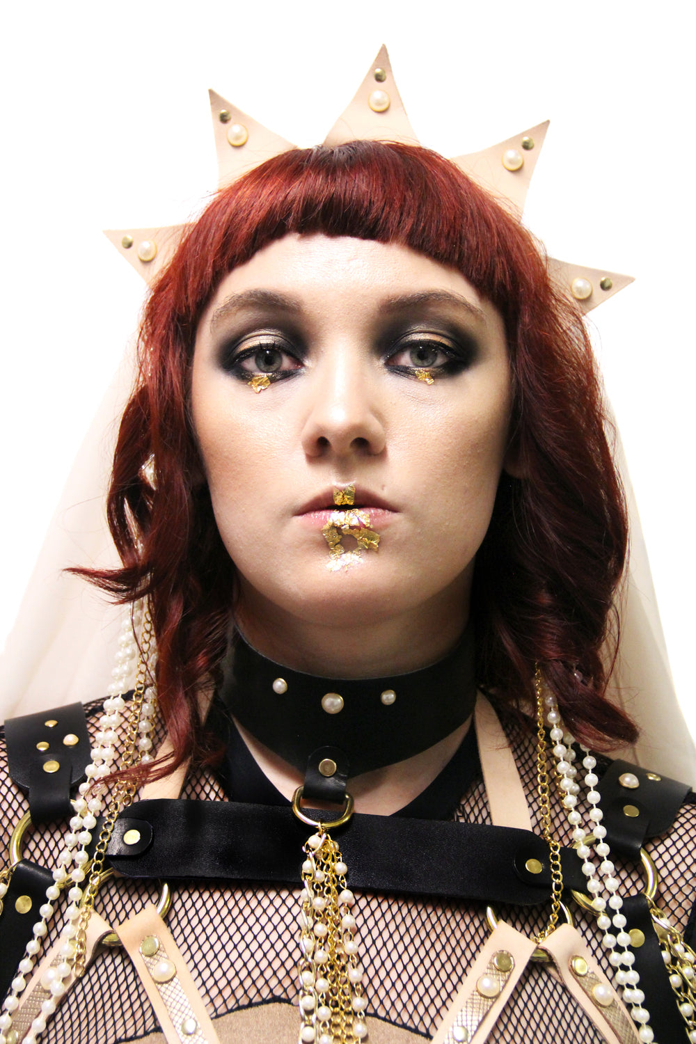 Heavily adorned model in gold foil makeup stares right into the camera. One of the accessories they are wearing is a pearl tassel choker.