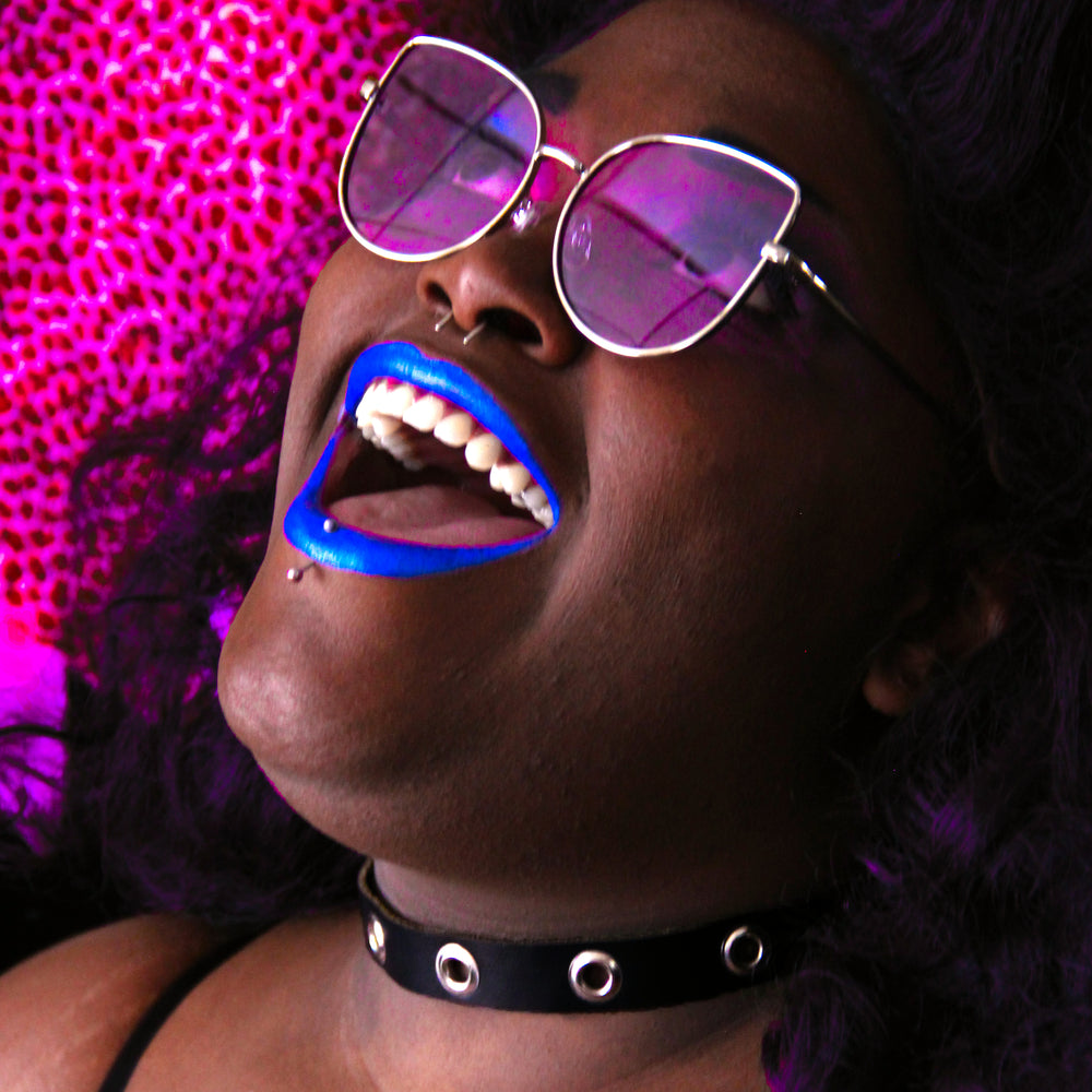 Blue-lipsticked model smiles widely while wearing shades with purple lenses and a black leather choker with silver grommets.