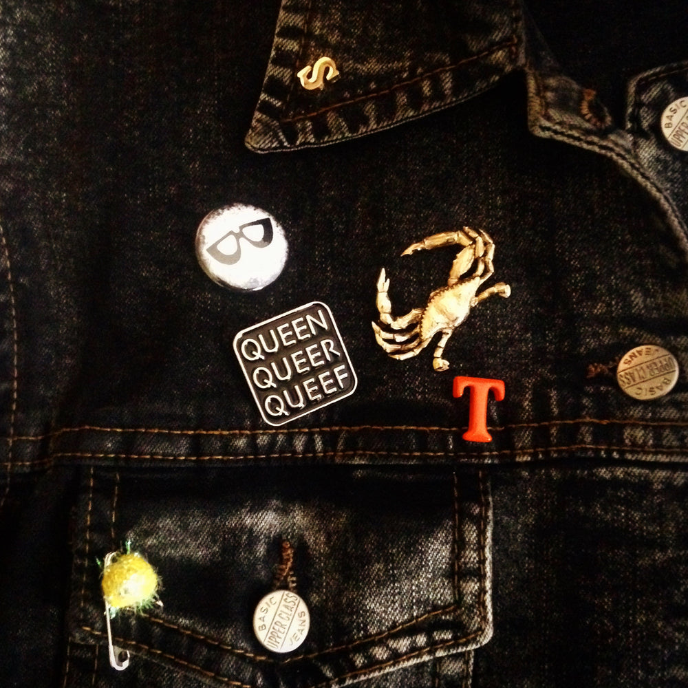 A black denim vest is adorned with many pins. Among them is a Q-Words pin, a vintage gold crab pin, some letters and others.