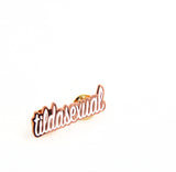 Close up of tildasexual pin. Copper metal outlines script lettering that reads “tildasexual” in white enamel.