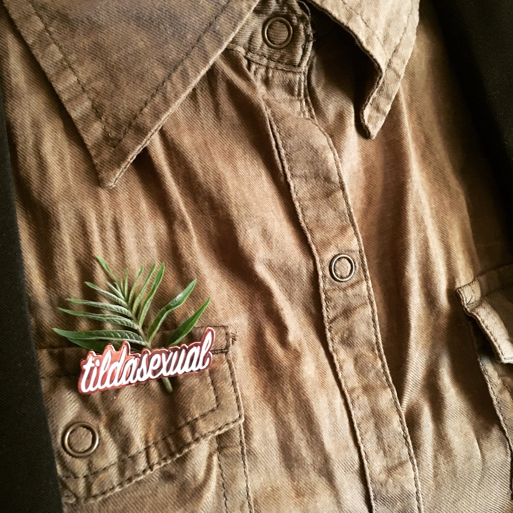 A small bit of foliage is pinned in place to an olive colored button up shirt with a tildasexual pin.