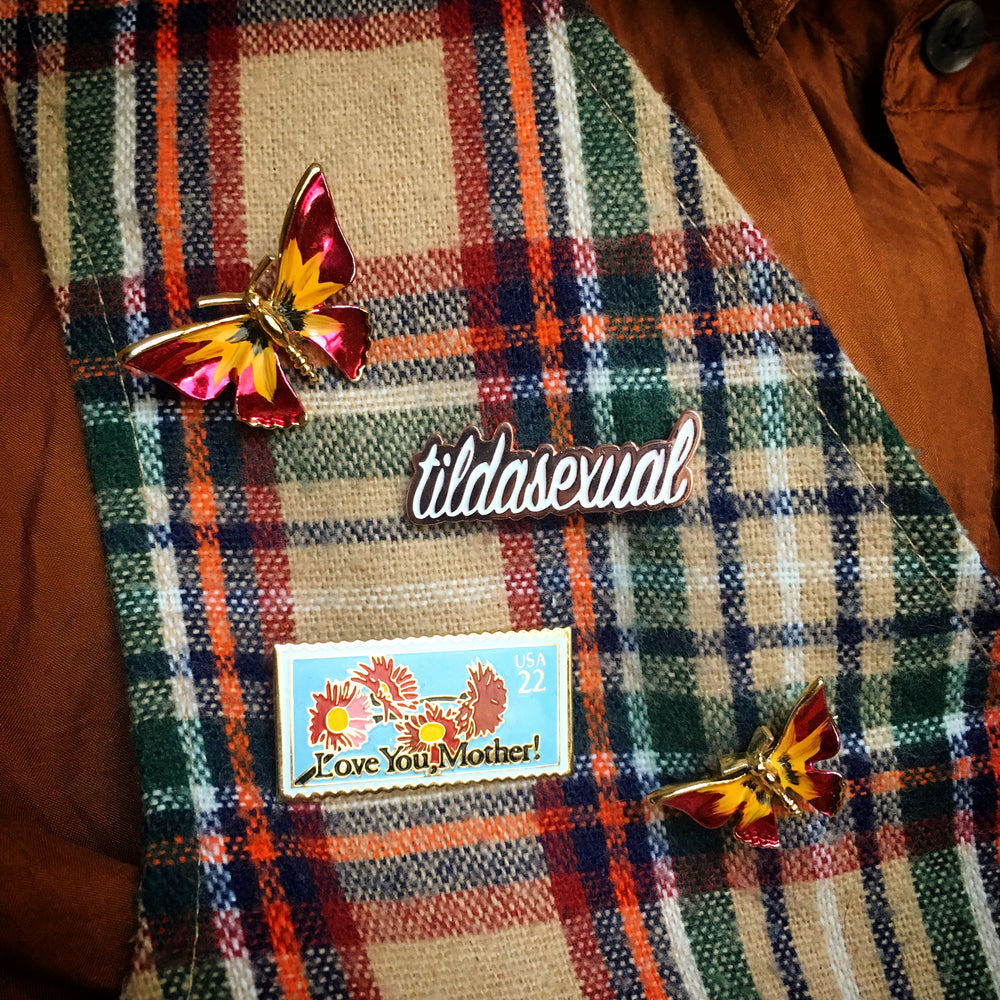 A plaid wool vest is adorned with two vintage butterfly pins, a tildasexual pin, and a postage stamp pin.