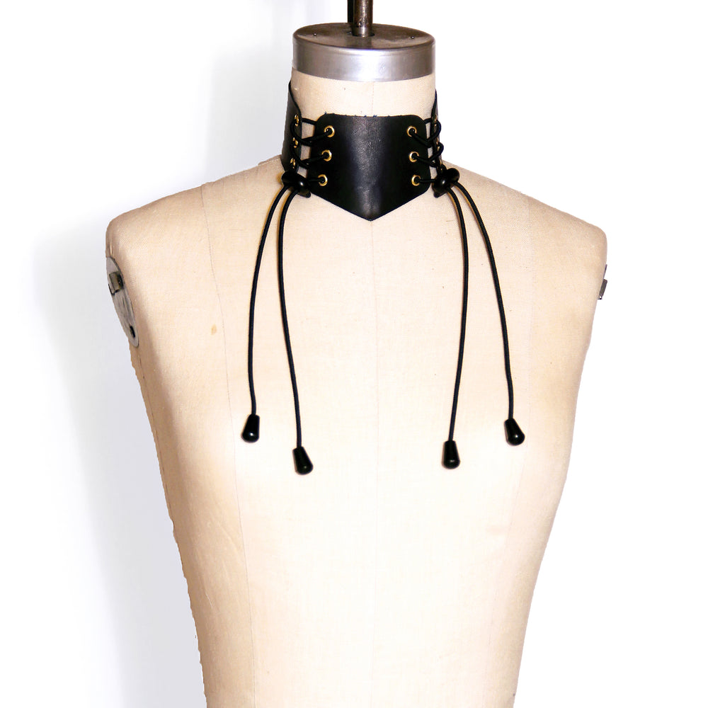 Wide black leather collar with two sets of brass eyelets laced with black cording. Shown on a dress form. 