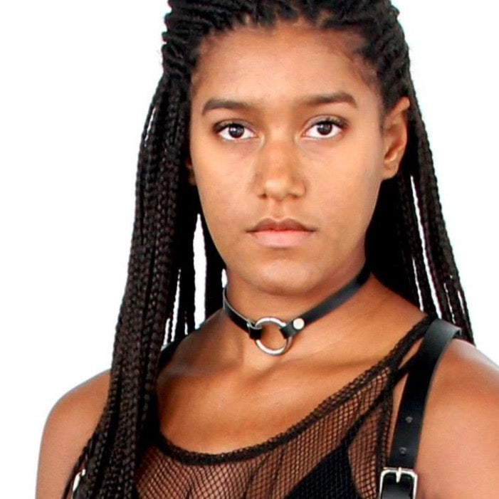 Model with long braids stares directly into camera while wearing a mesh top, a chest harness, and a thin black leather o-ring choker.