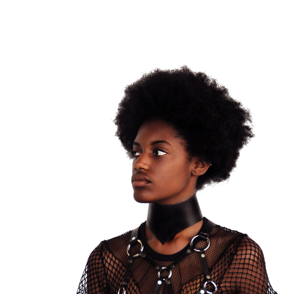 Model in mesh top and leather harness wears a leather posture collar while looking off to the side.
