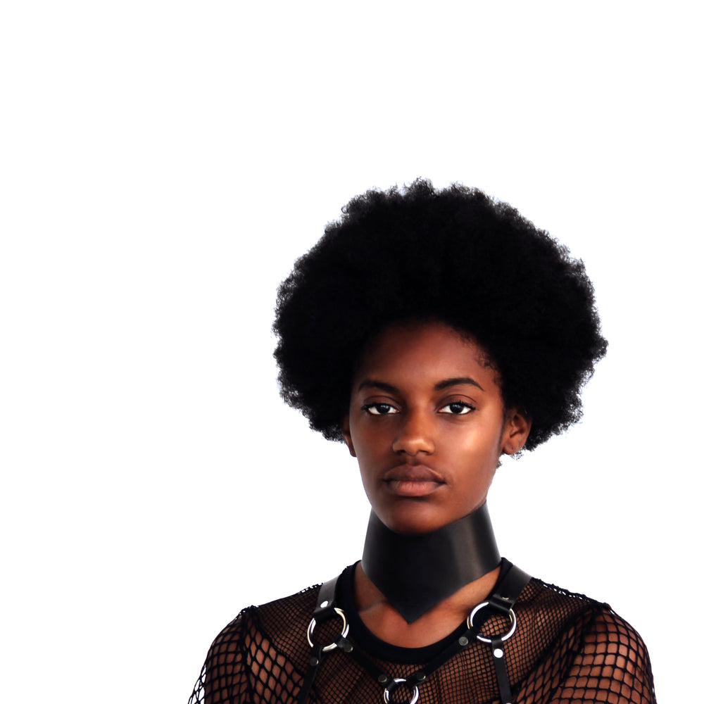 Model with an afro stares directly into the camera while wearing a posture collar, mesh top and leather harness.