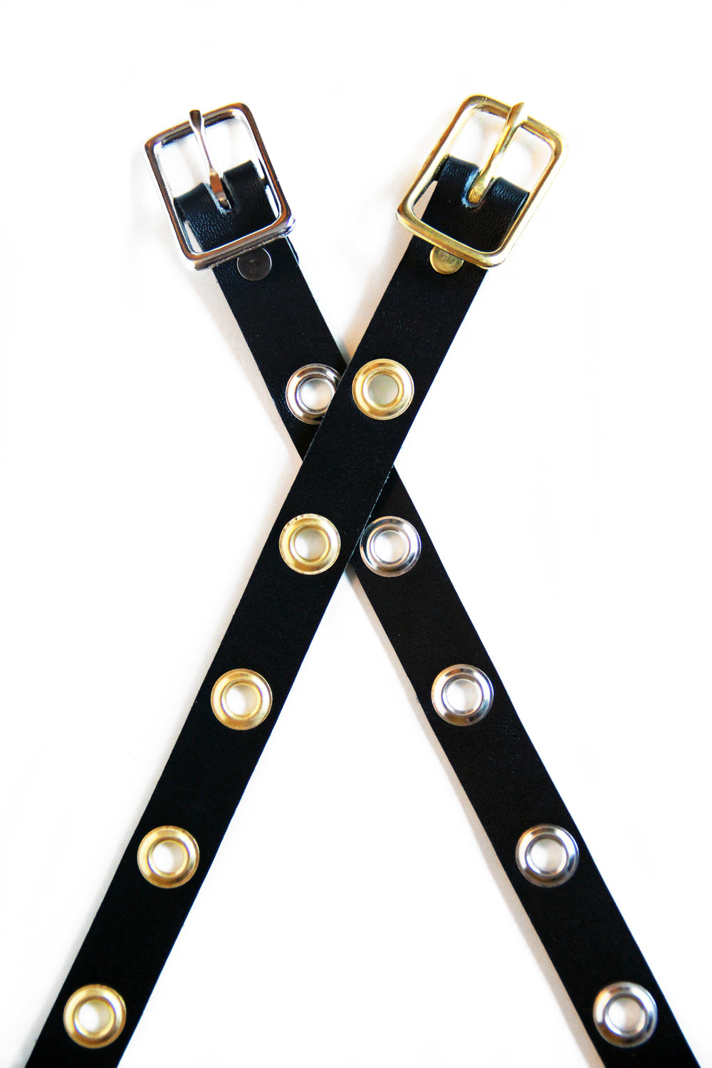 Two leather choker laid flat in the shape of an 'X.' One choker has silver grommets an the other has brass.
