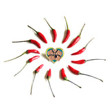 One leather daddy pin lays on a white background with small red peppers arranged in a swirling circle surrounding it.