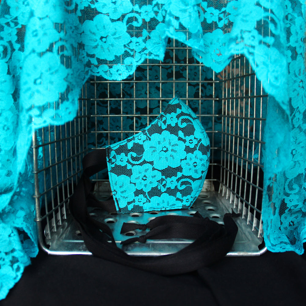 Teal lace face mask in a wire cage draped with more teal lace fabric. Black cotton ties of masks showing.