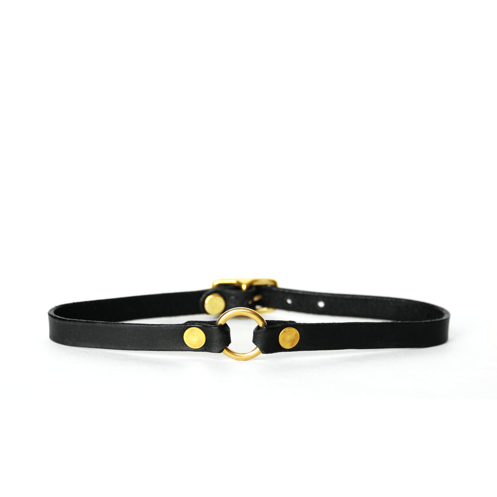 A thin black leather choker with a tiny brass o-ring at center front sits on a white surface with a white background.