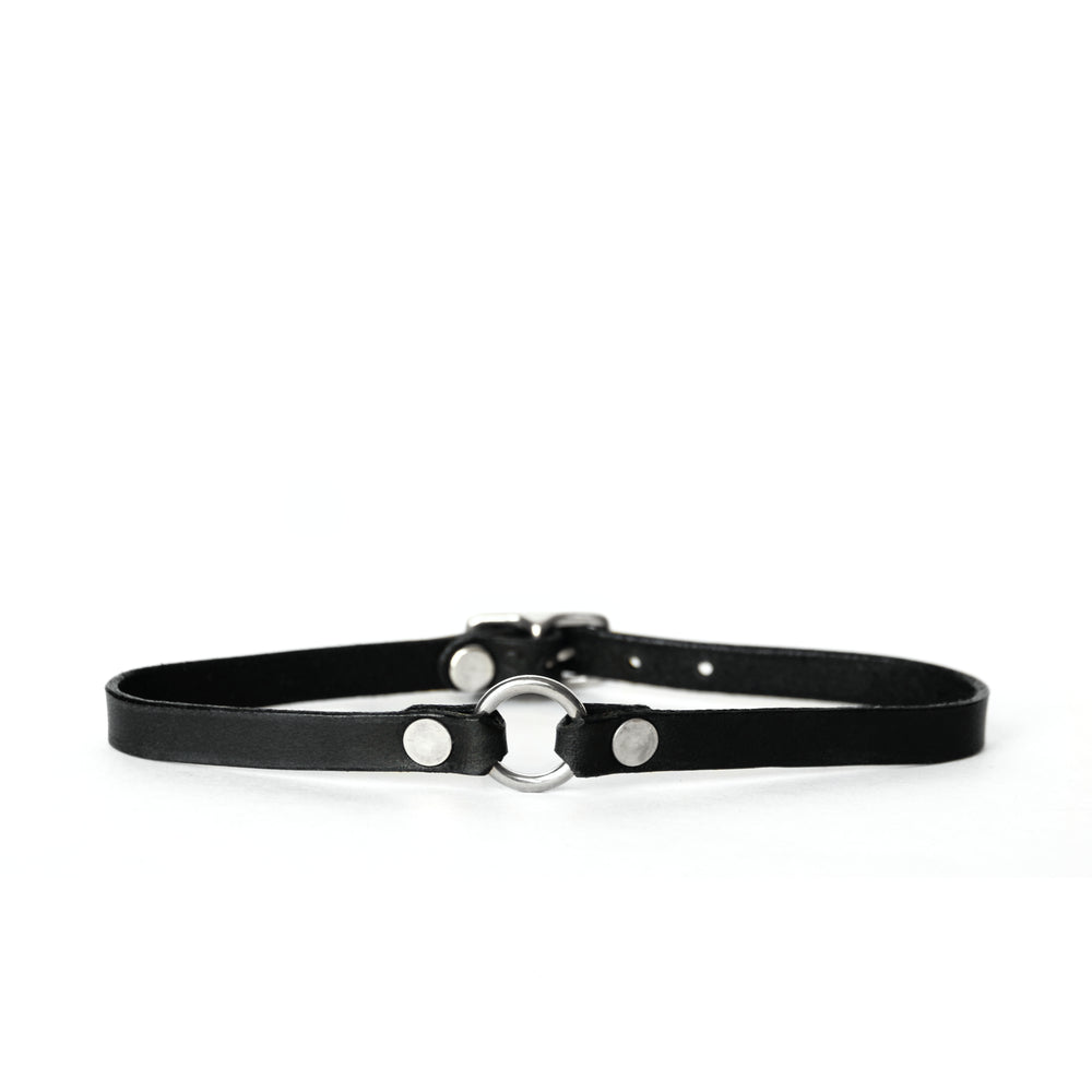 A thin black leather choker with a tiny silver o-ring at center front sits on a white surface with a white background.