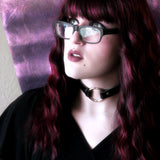 A bespectacled model stands in a glowing light while wearing lipstick, a a black dress, and a black o-ring choker. 