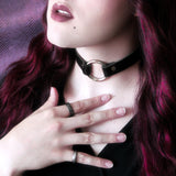 Lipsticked model in soft pink light caresses their nape of neck near a black choker with a silver o-ring. 