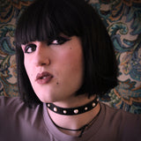 Model with a black bob wig wears a black leather collar studded with pearls against a brocade background.