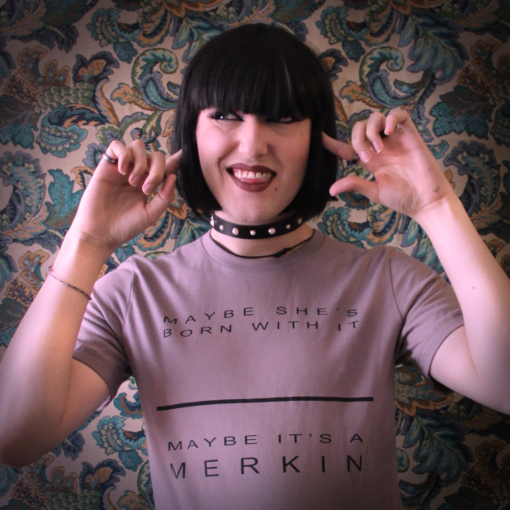Model smiles with tongue out while wearing a tee and a leather and pearl collar. Tee reads "maybe she's born with it, maybe it's a merkin."