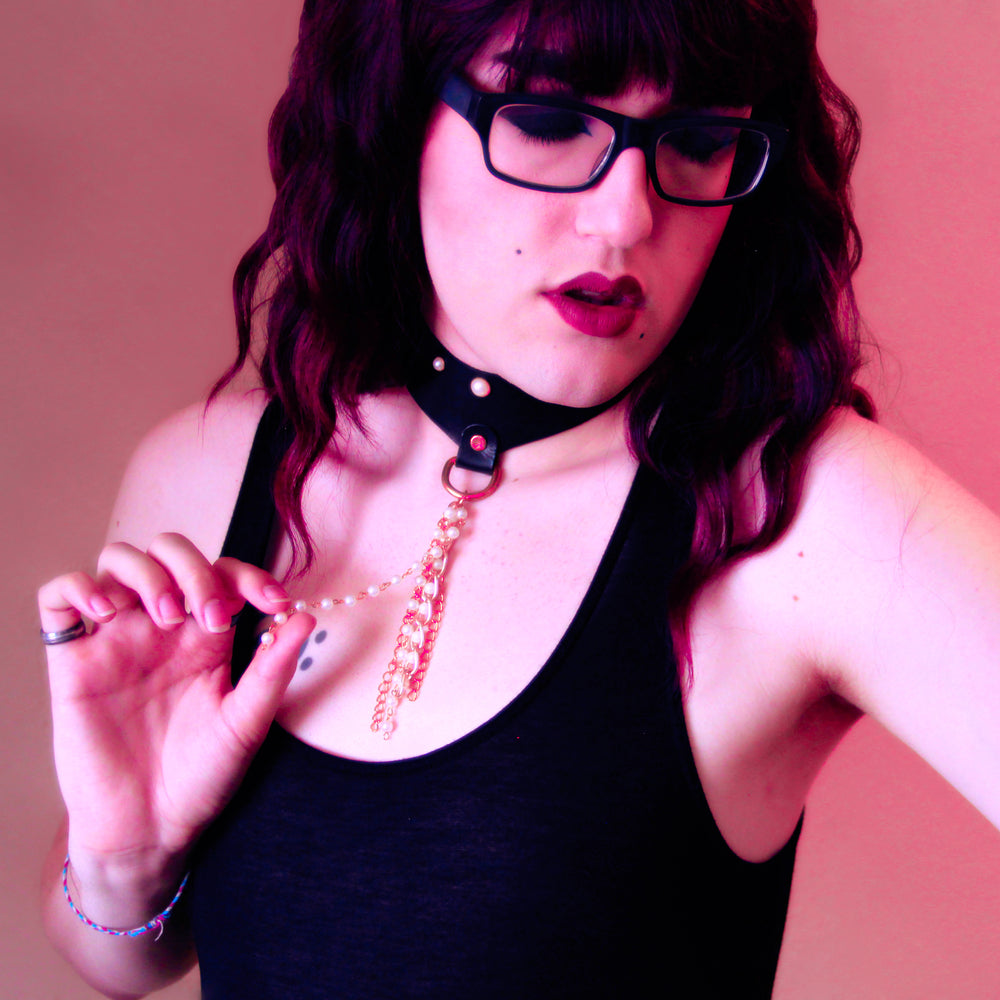 Lipsticked model bathed in pink toned light fondles the pearl strands in the tassel of the choker they are wearing. 