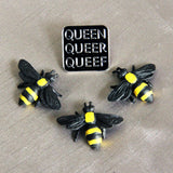A trio of plastic toy bees stand before a Q-words pin on a silver-toned fabric background.