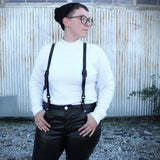 Model in a white mock turtleneck, black stocking cap and black faux leather pants is accessorized with black suspenders in front of a stark metal building.