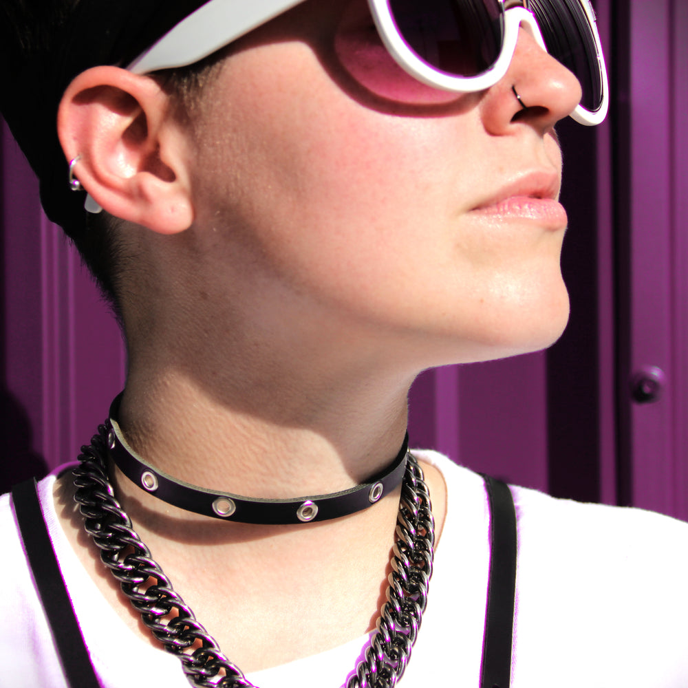 Model faces the sunshine in sunglasses, a thin black leather choker with silver grommets, a chain and a chest harness.