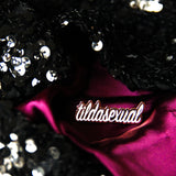 A tildasexual pin is laid upon fuchsia satin fabric and framed by black sequined fabric.