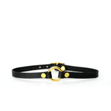 A thin black leather choker with a small brass o-ring at center front sits on a white surface with a white background.